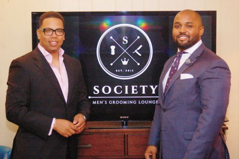 GROOMED FOR SUCCESS—Society owners K. Chase Patterson, left, and Ryan Norman pose in front of their logo shown on the HD screen in the grooming lounge’s downstairs sitting room. (Photo by Rossano P. Stewart)